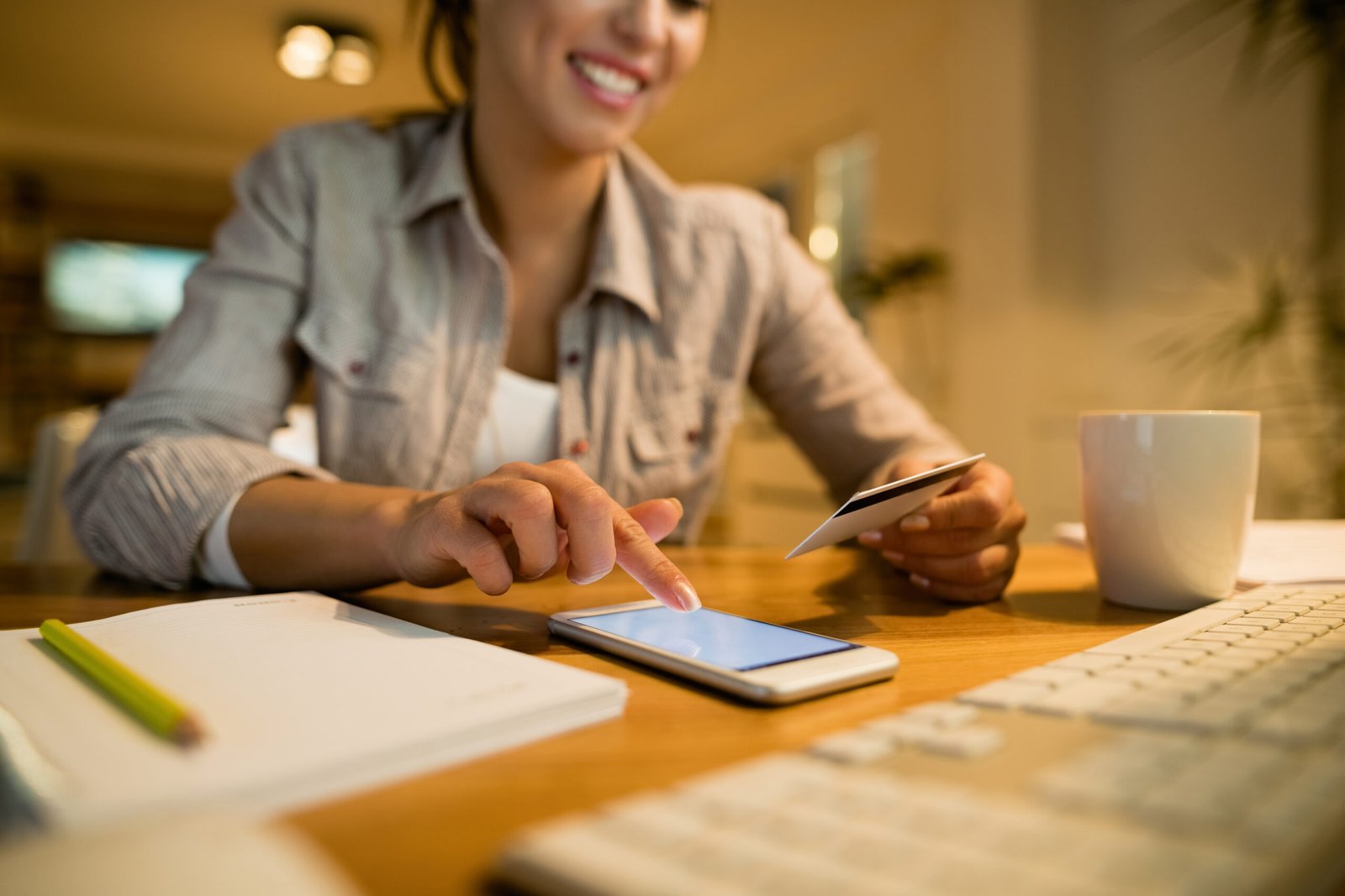 Mulher sorrindo enquanto utiliza um smartphone e segura um cartão de crédito sobre a mesa, com caderno, teclado e caneca ao redor, representando compra online, pagamento digital e uso de tecnologia no dia a dia. Imagem de capa para o blogpost Split Payment na Reforma Tributária 2026