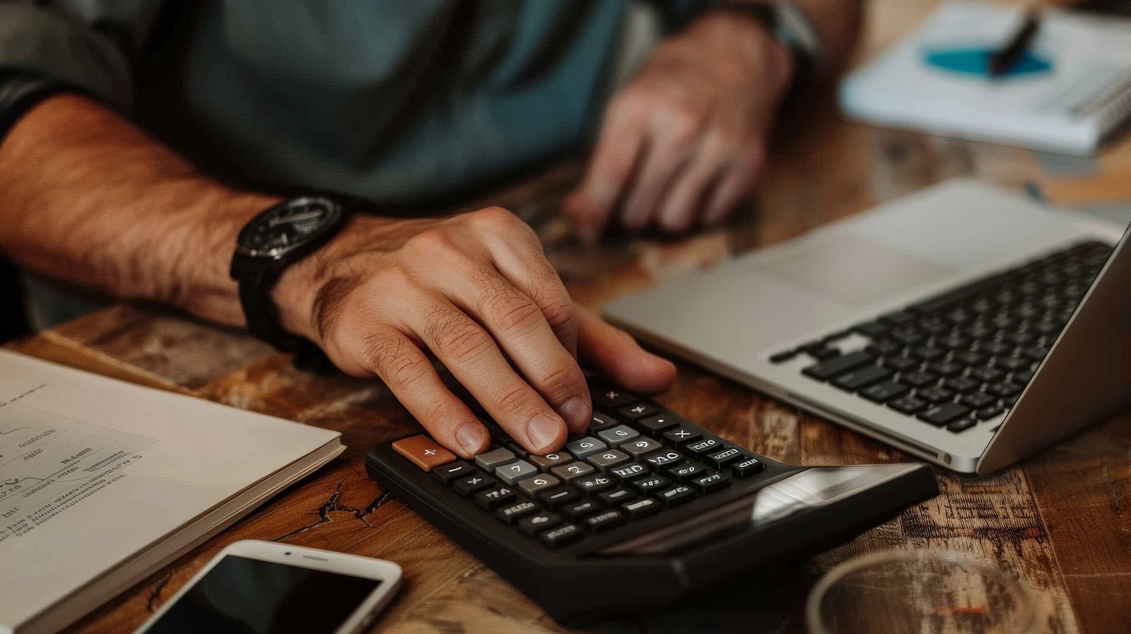Pessoa utilizando uma calculadora financeira sobre uma mesa de madeira, ao lado de um laptop, documentos e um smartphone, representando planejamento financeiro, análise de custos e controle de investimentos. Imagem de capa para o blogpost Os Novos Impostos da Reforma Tributária e como estar preparado para as mudanças